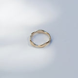 A single gold-toned ring with a wavy, organic textured band, photographed on a light gray surface with soft, dappled shadows.