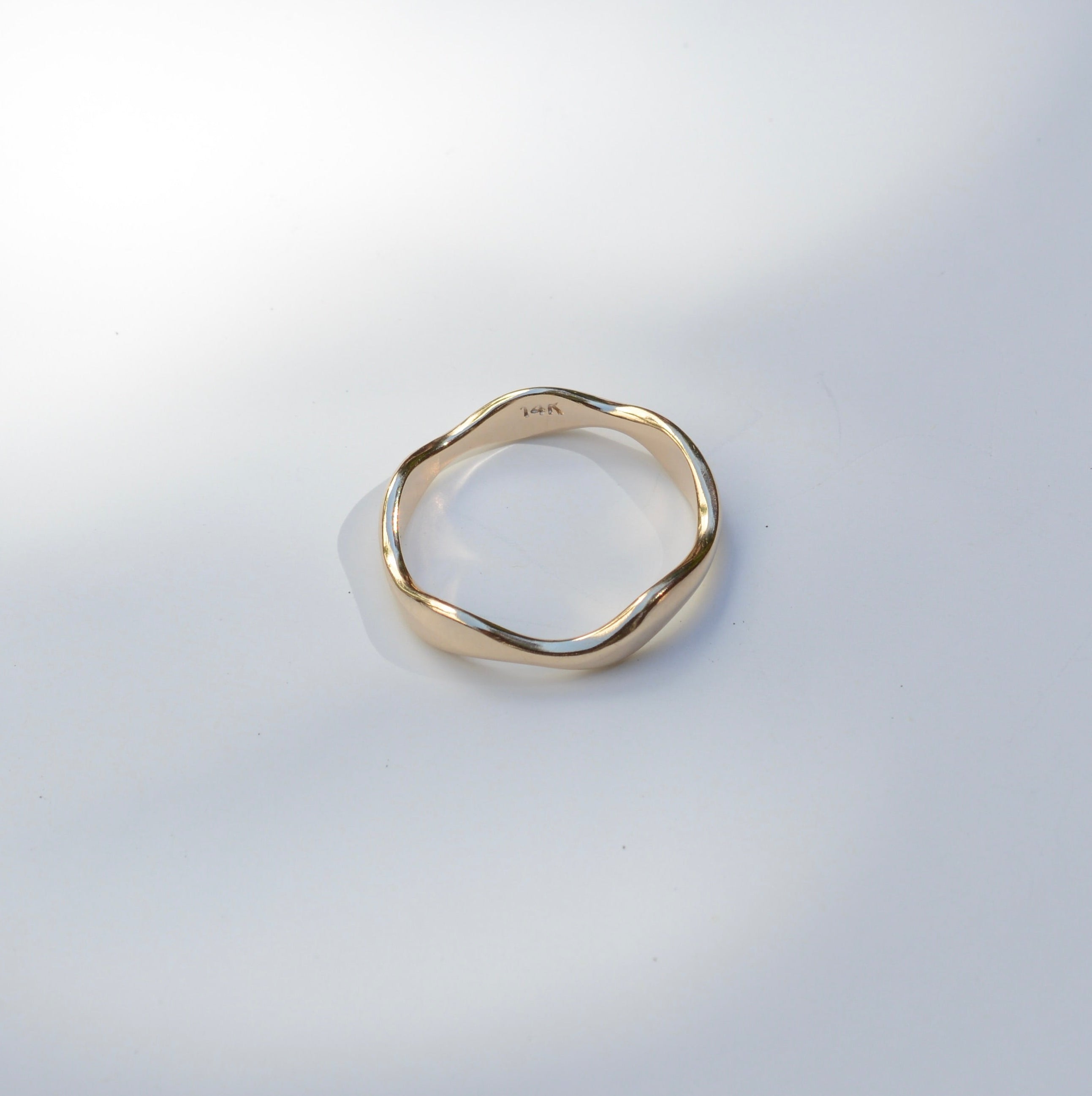 A single gold-toned ring with an organic, wavy, irregular shape, photographed on a light gray surface with soft, artistic shadows.