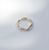 A single gold-toned ring with an organic, wavy, irregular shape, photographed on a light gray surface with soft, artistic shadows.
