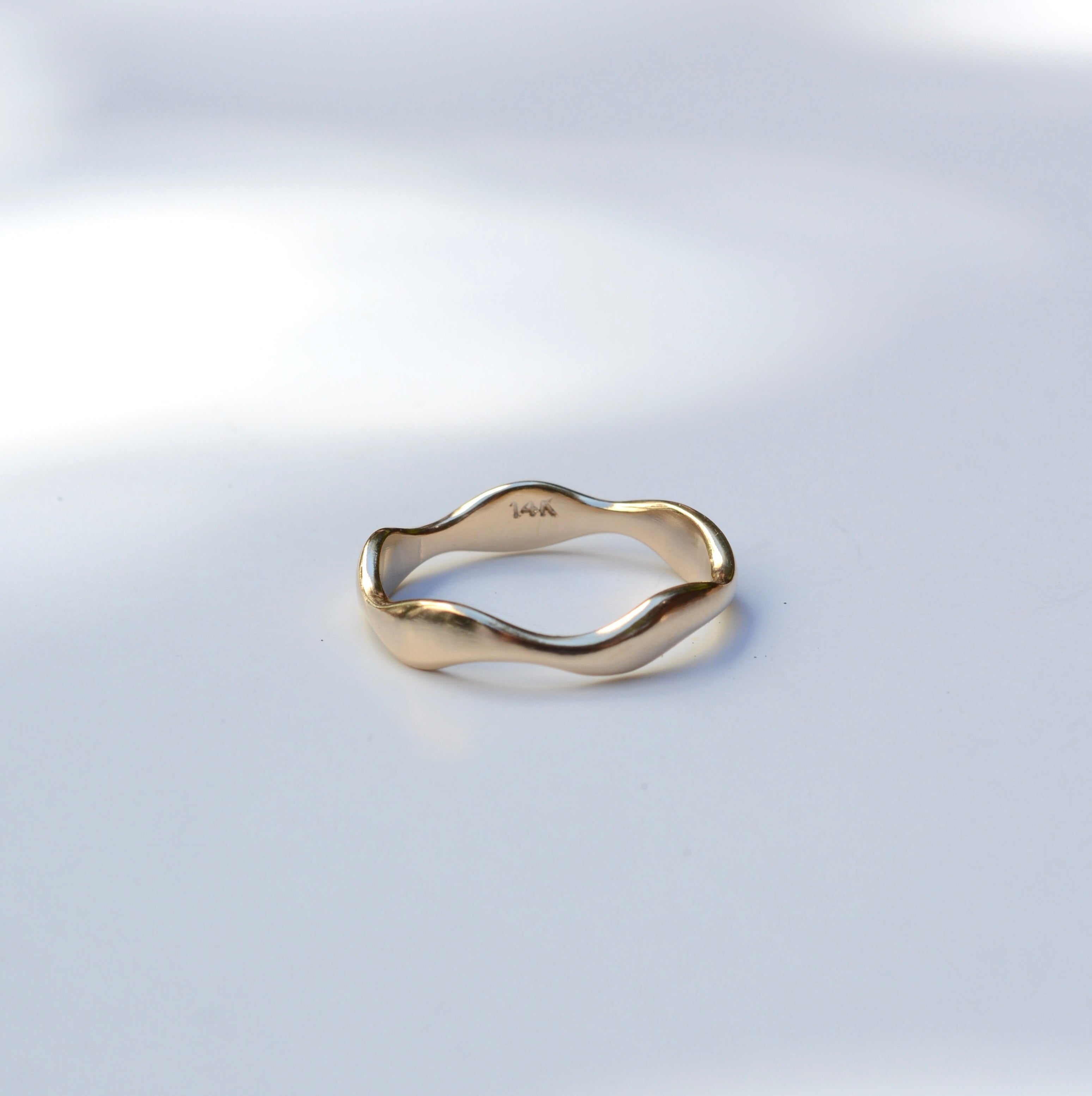 A gold wavy band ring with a polished finish, resting on a light gray surface with soft, dappled shadows.