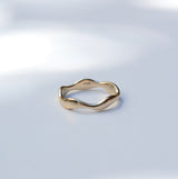 A gold wavy band ring with a polished finish, resting on a light gray surface with soft, dappled shadows.