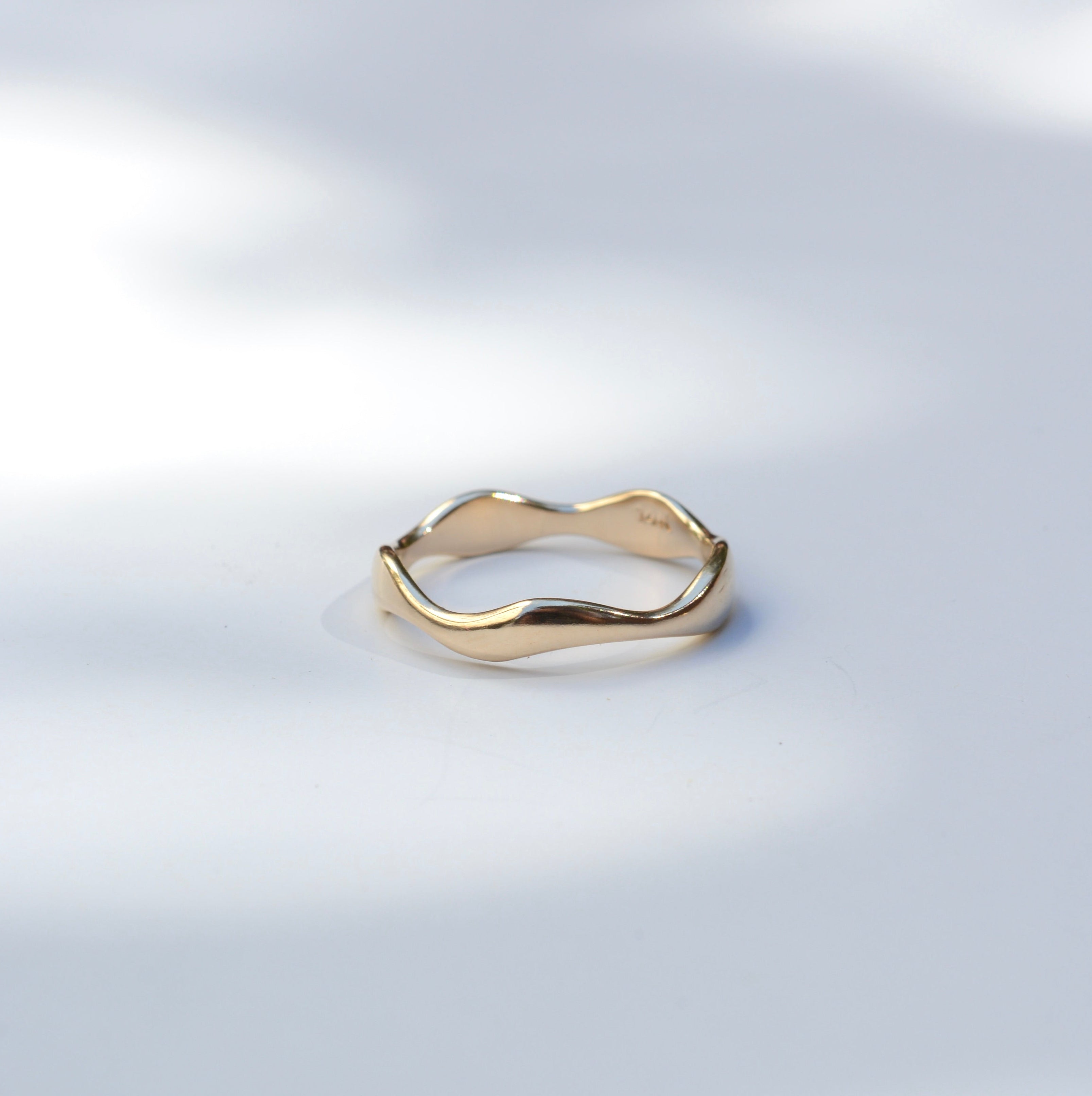 A slim, wavy gold ring with a polished finish, resting on a light gray surface with soft, dappled shadows.