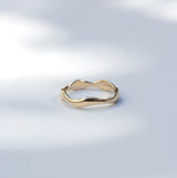 A slim, wavy gold ring with a polished finish, resting on a light gray surface with soft, dappled shadows.