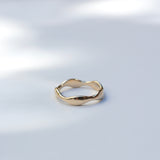 A single gold ring with an organic, wavy, undulating band design, photographed on a light gray surface with soft, dappled shadows.