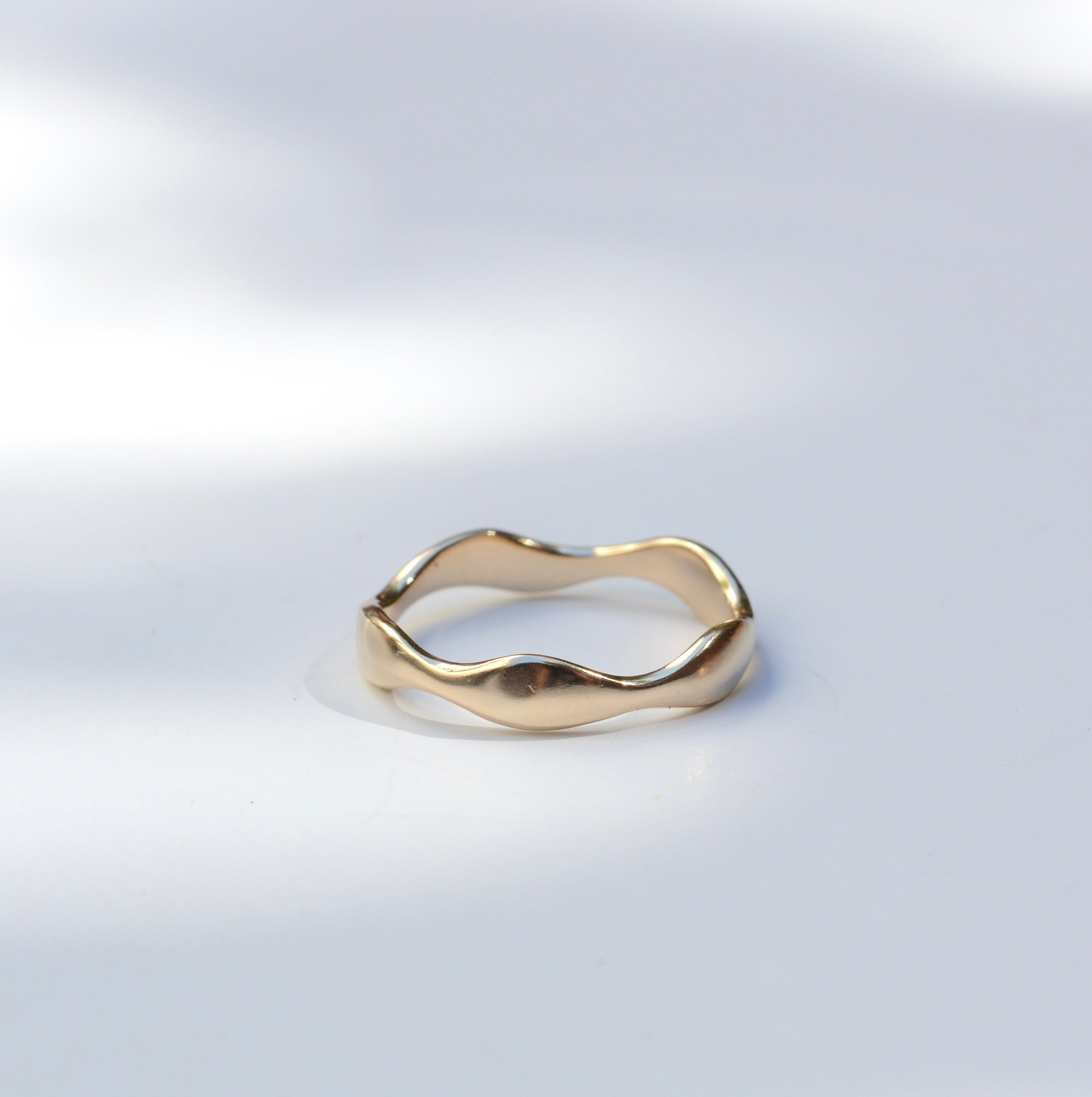 A single gold ring with an organic, wavy, undulating band design, photographed on a light gray surface with soft, artistic shadows.