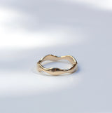 A single gold ring with an organic, wavy, undulating band design, photographed on a light gray surface with soft, artistic shadows.