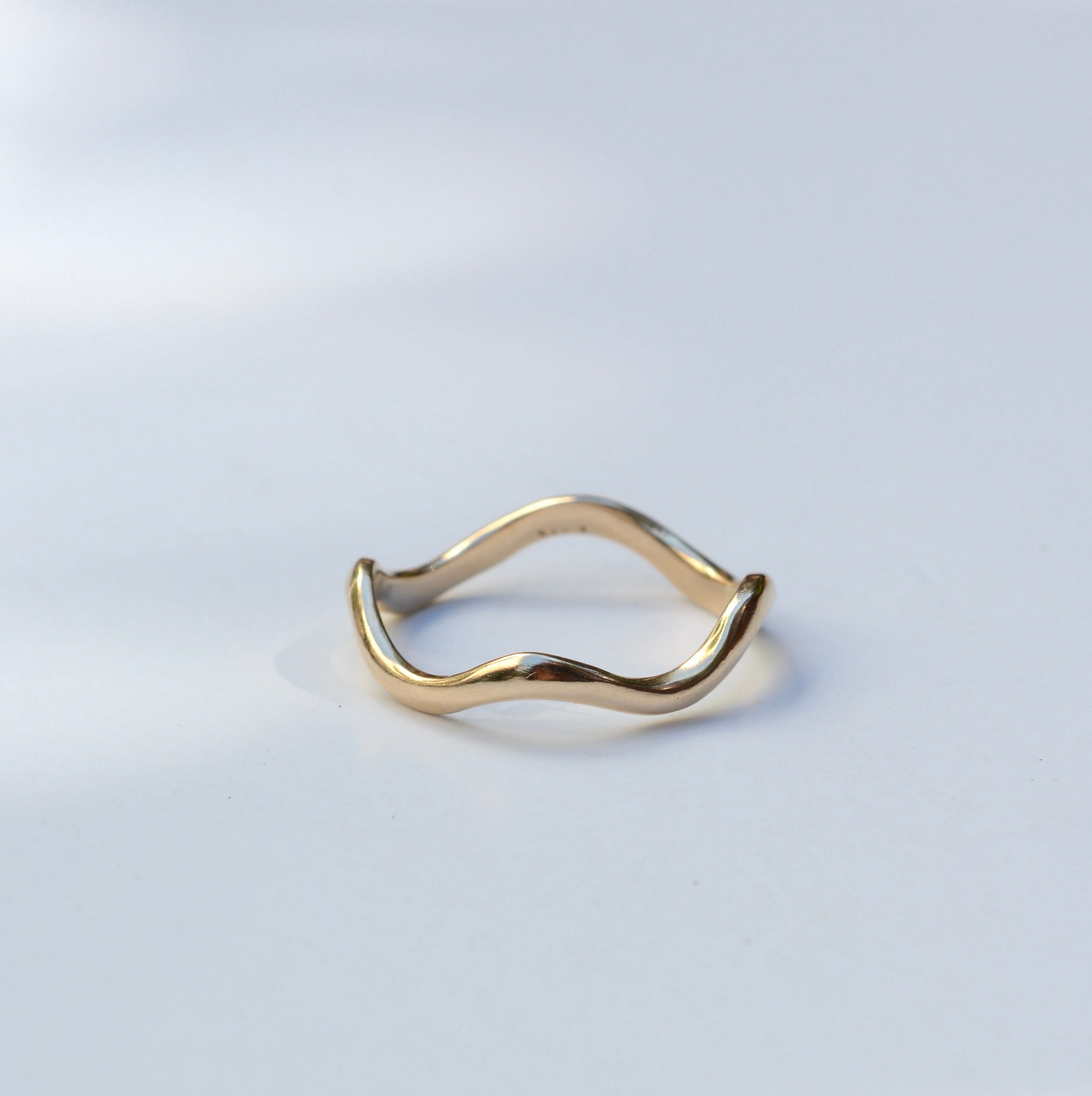 A thin, wavy gold ring with a unique undulating band, photographed on a clean white surface with soft, natural-looking shadows.