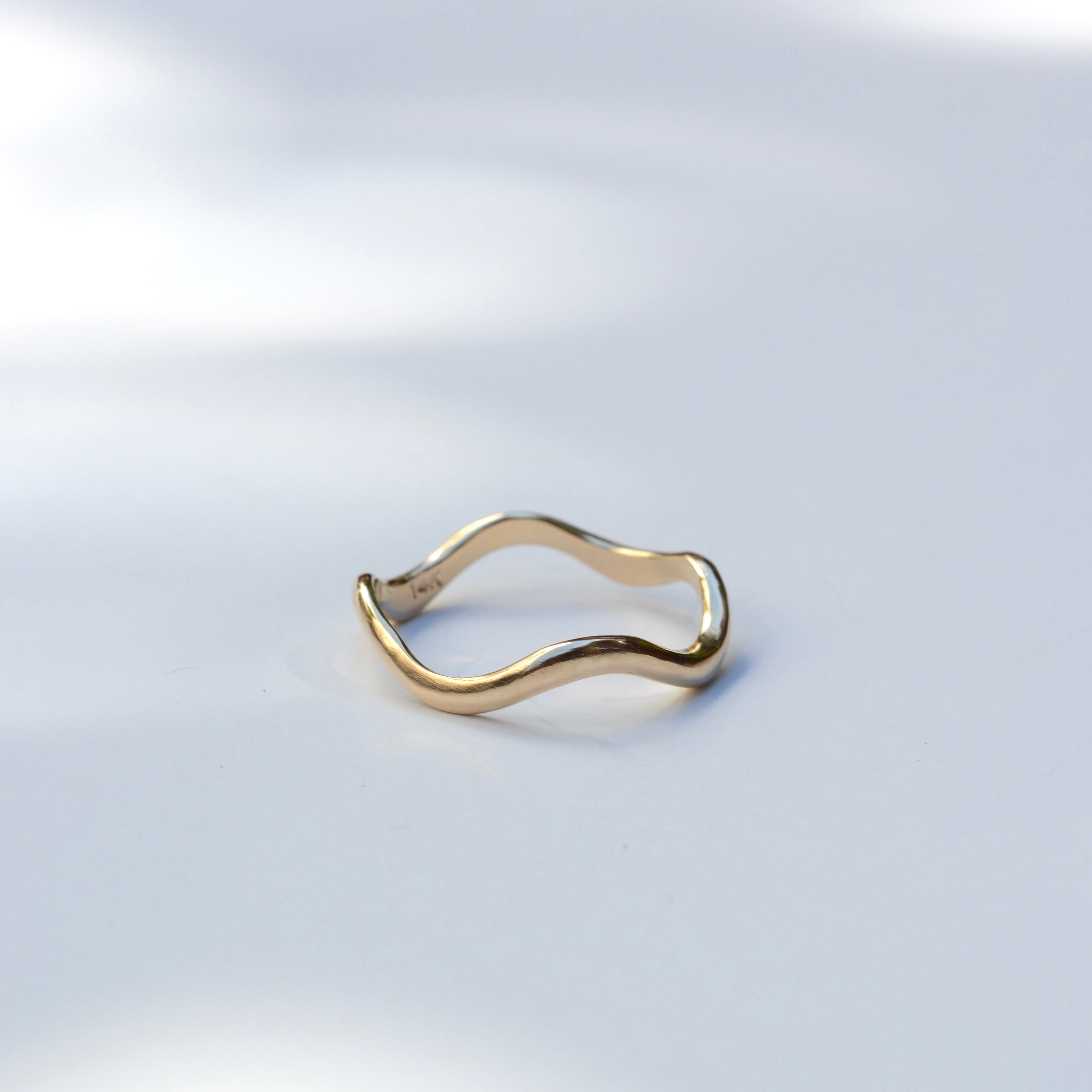A thin, wavy gold ring with a polished finish, displayed on a clean white surface with soft, natural leaf shadows.