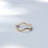 A thin, wavy gold ring with a polished finish, displayed on a clean white surface with soft, natural leaf shadows.