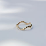 A thin, wavy gold ring with a polished finish, resting on a white surface with soft, natural leaf shadows.
