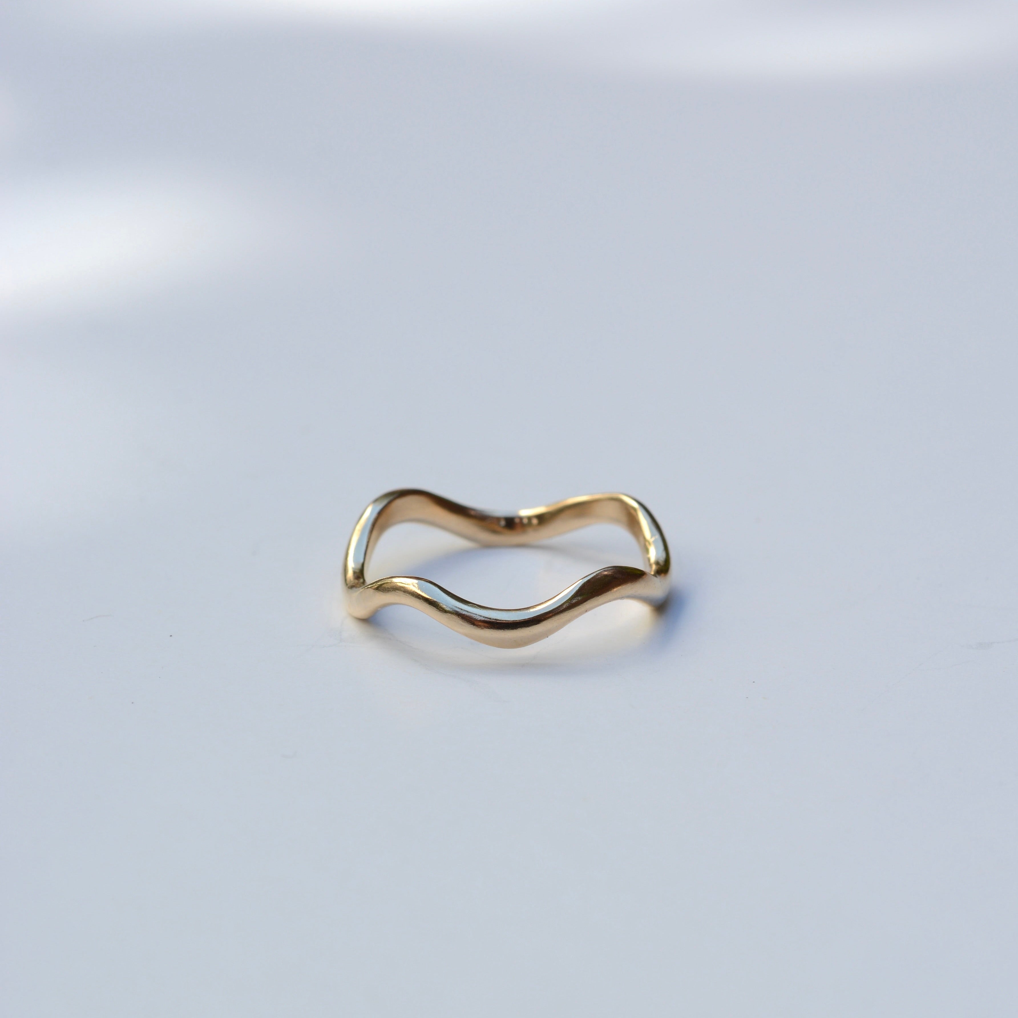 A thin, wavy gold band ring photographed on a clean white surface with soft shadows.