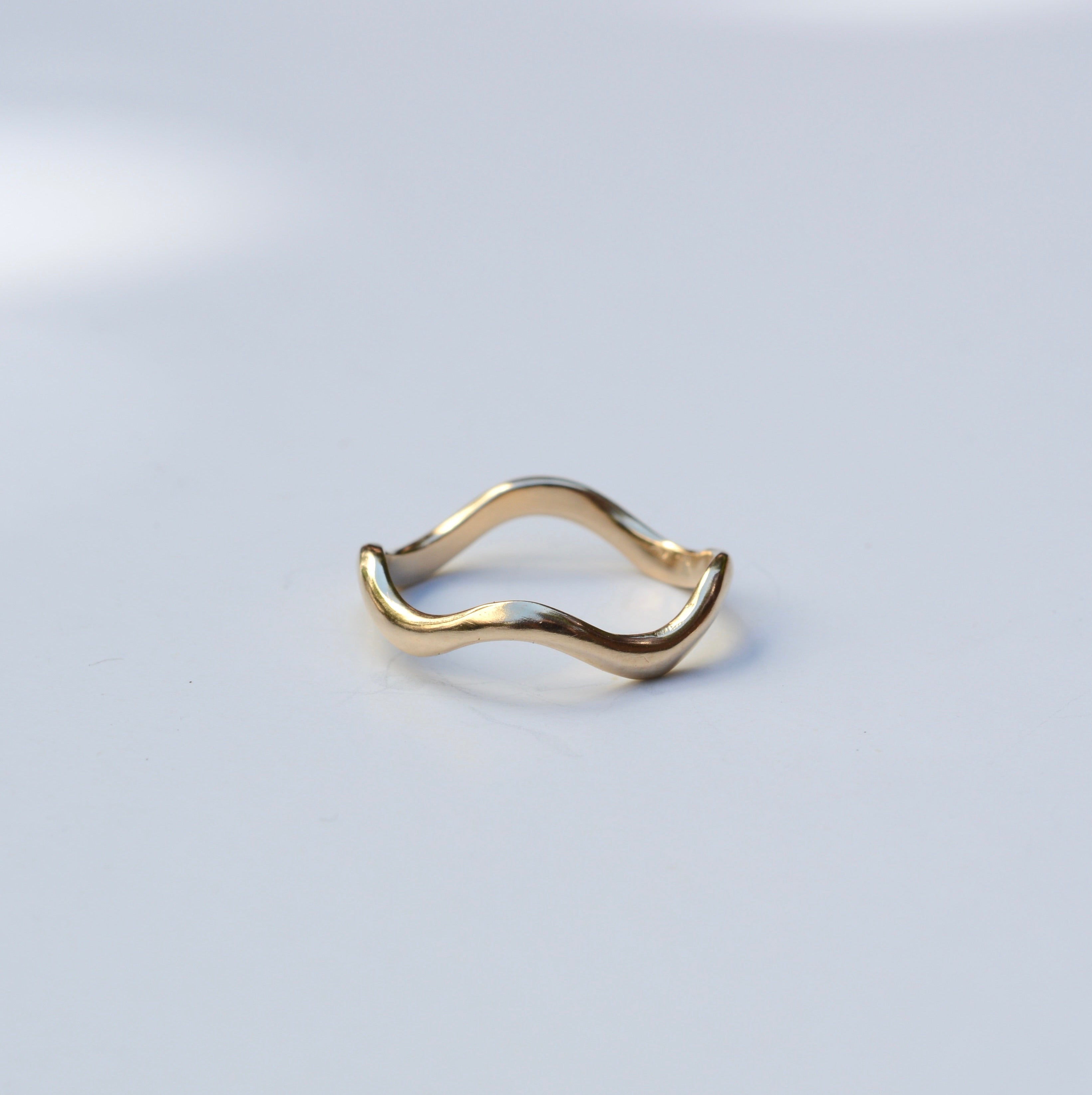 A single gold ring with a wavy, undulating band, photographed on a clean, light gray surface with soft shadows.