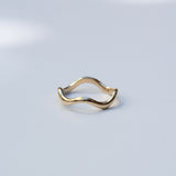 A single gold ring with a wavy, undulating band, photographed on a clean, light gray surface with soft shadows.
