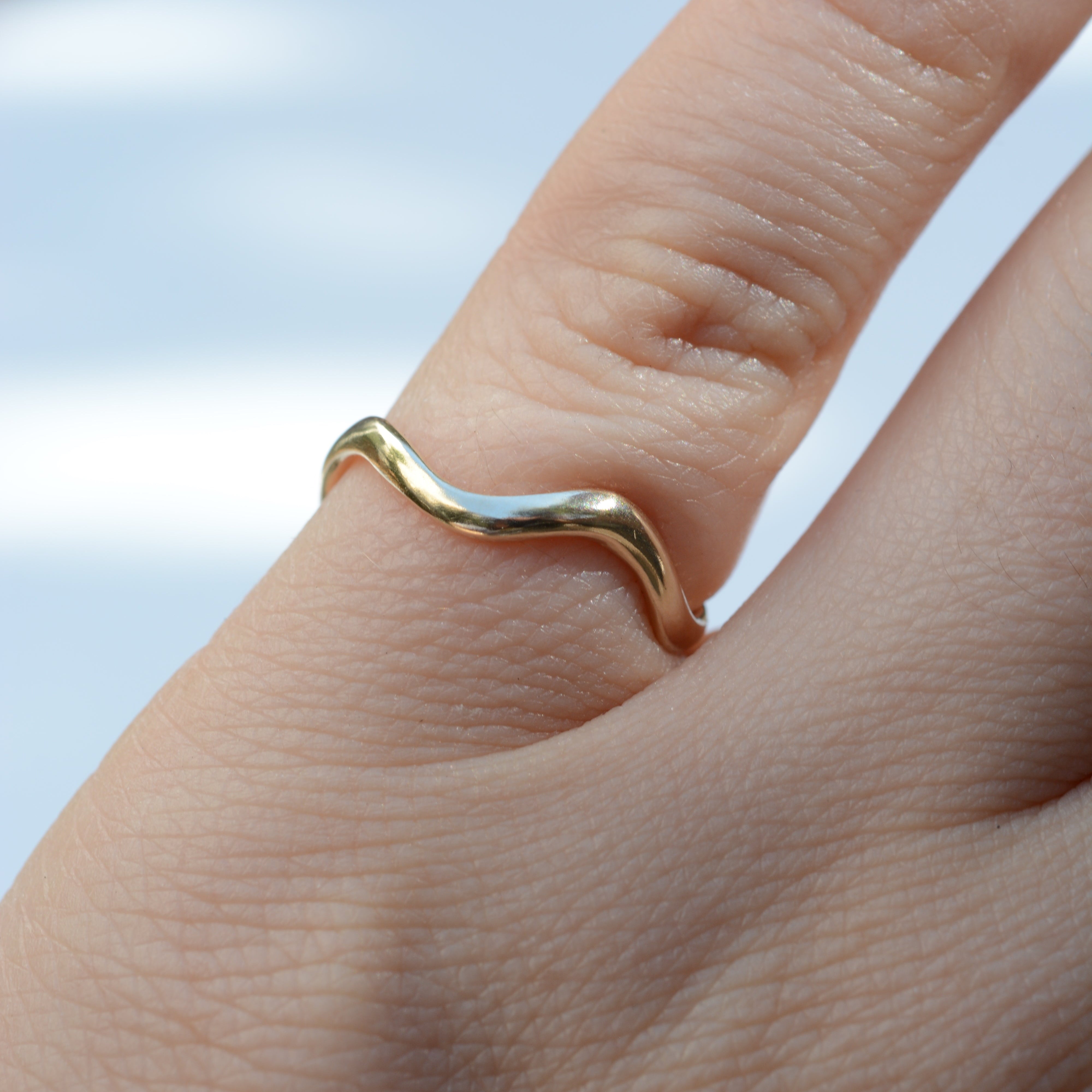 A close-up of a hand wearing a thin, wavy gold ring on the ring finger against a softly blurred light background.