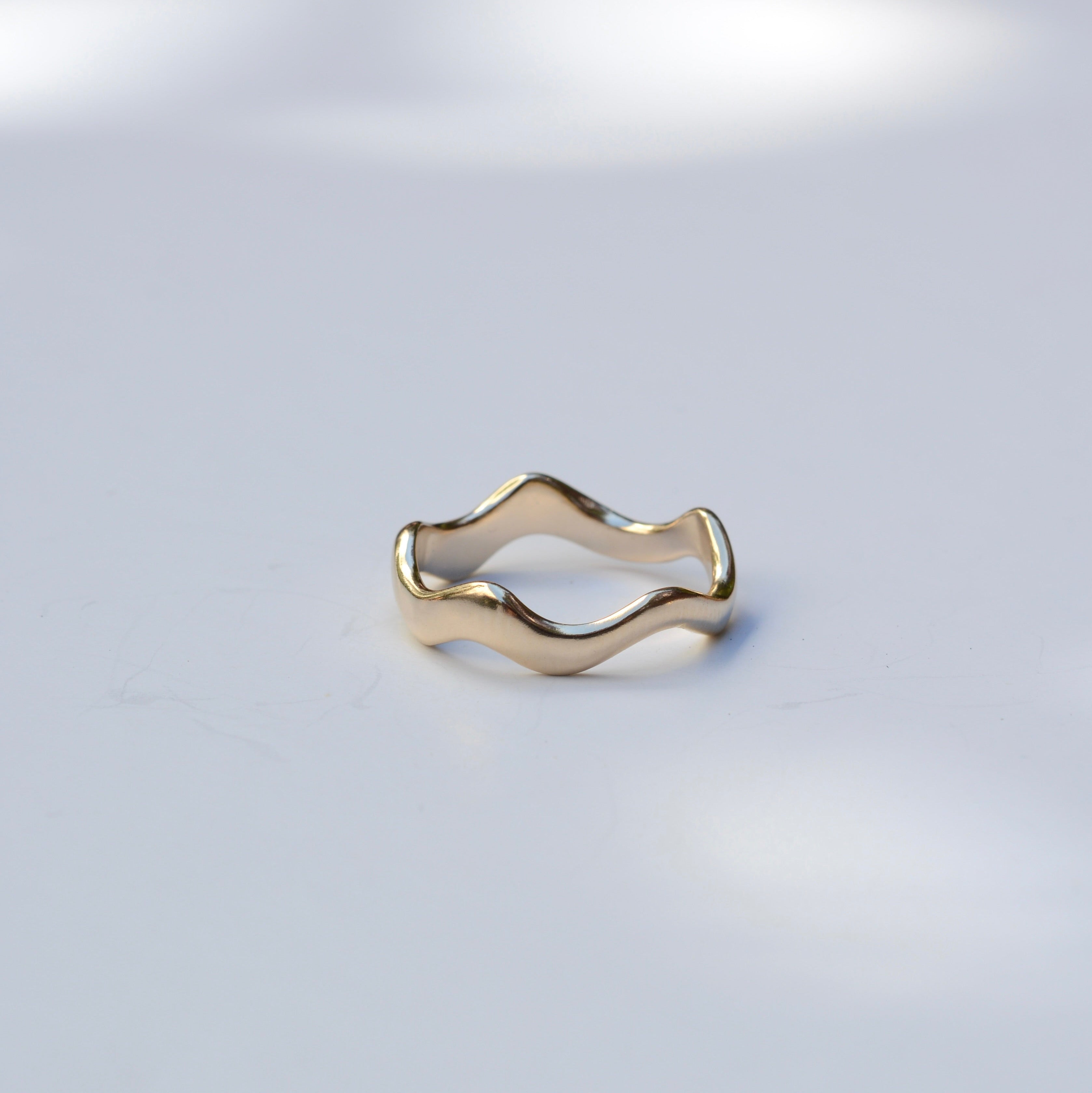 A gold wavy open-band ring resting on a light gray surface with soft, dappled shadows.