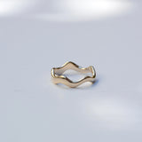A gold wavy open-band ring resting on a light gray surface with soft, dappled shadows.