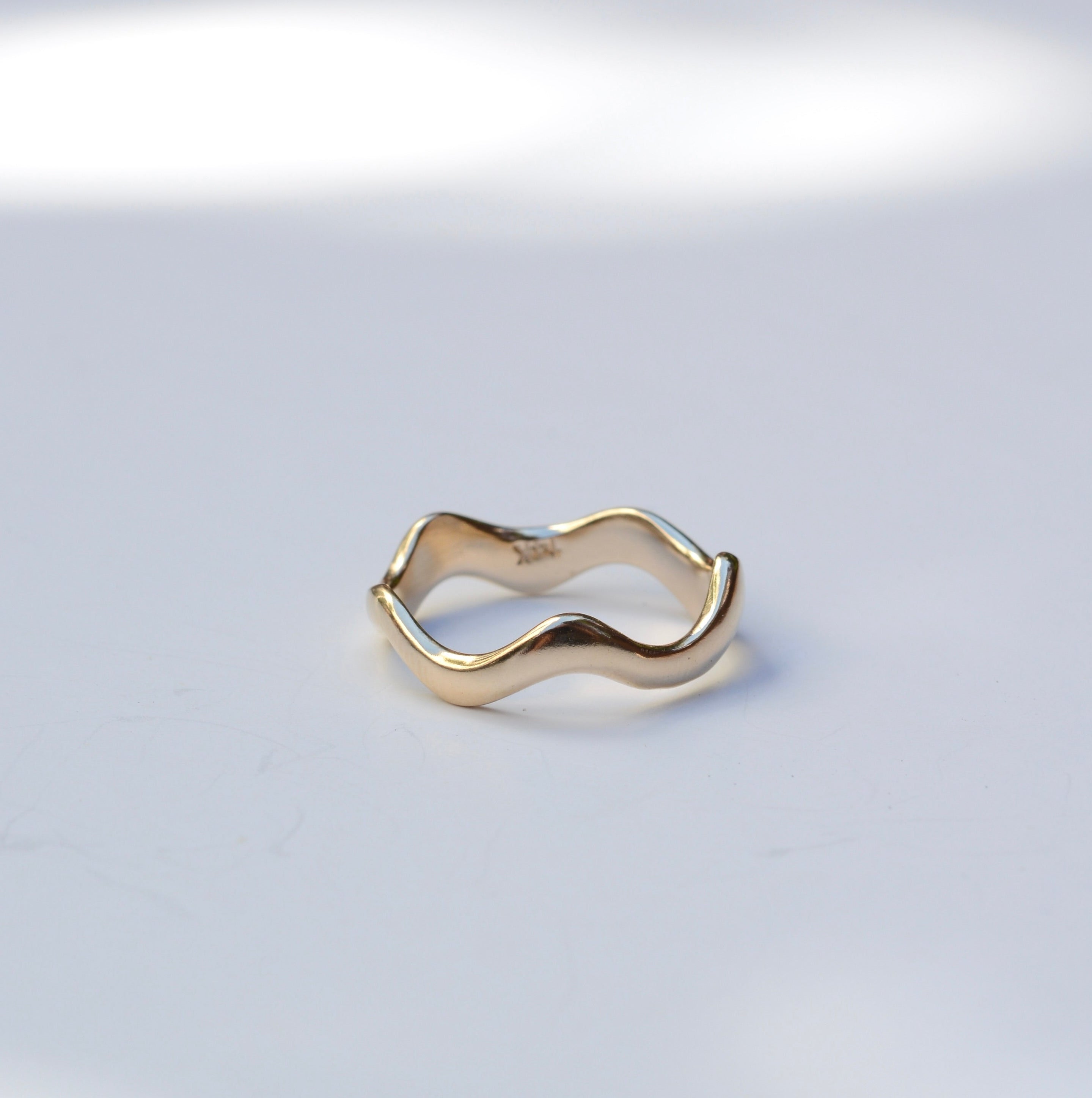 A gold wavy open ring with a minimalist, fluid design, photographed on a light gray surface with soft, natural-looking shadows.