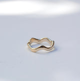 A gold wavy open ring with a minimalist, fluid design, photographed on a light gray surface with soft, natural-looking shadows.