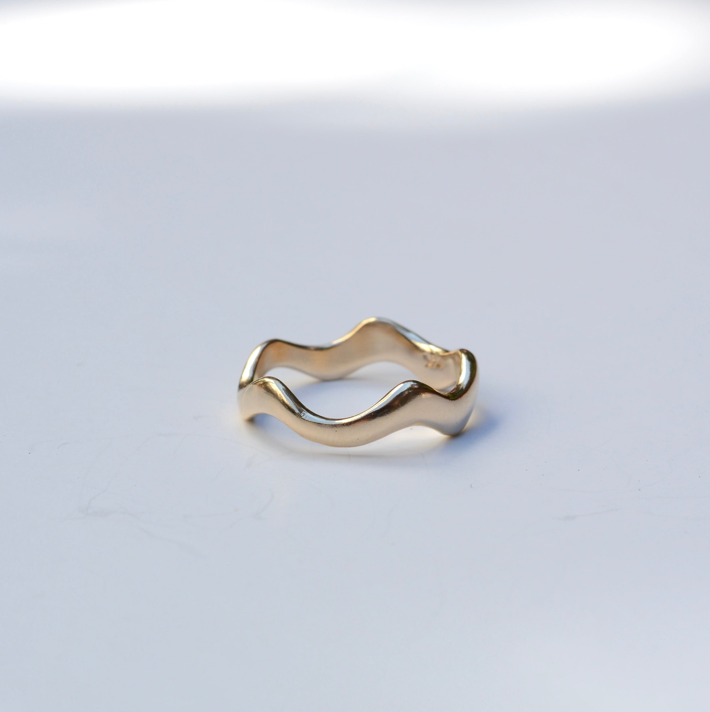 A single gold wavy band ring photographed on a clean white surface with soft natural shadows.