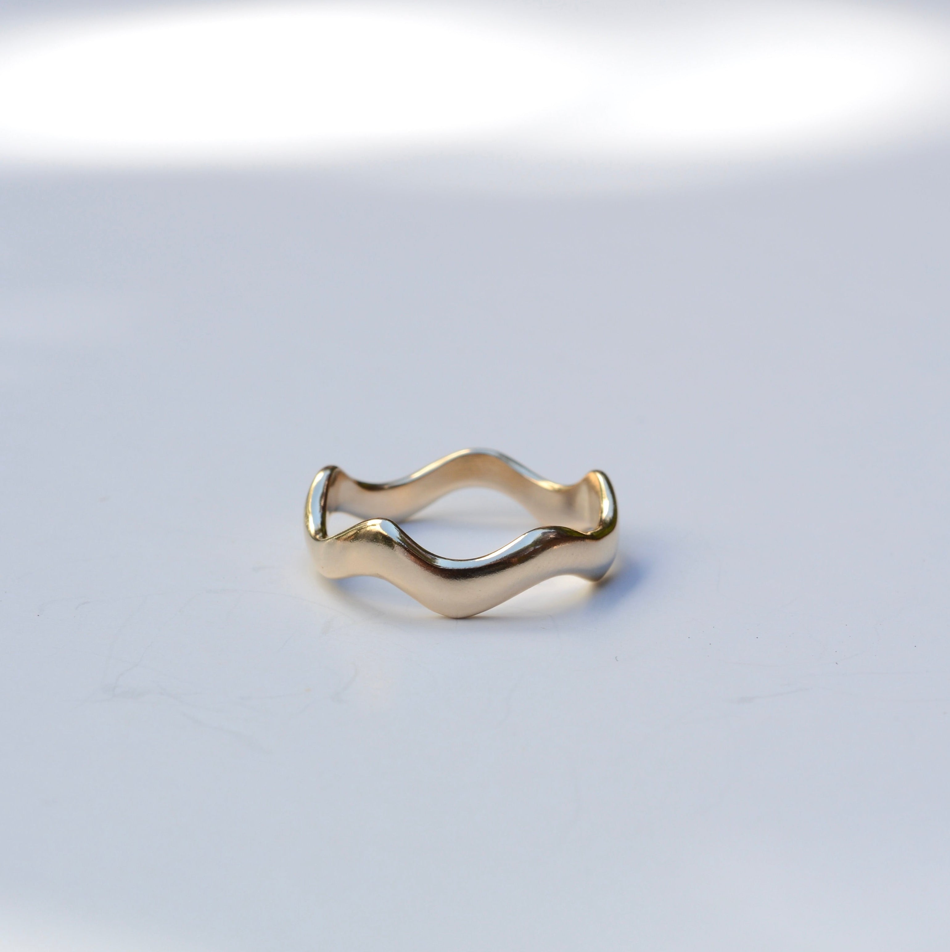 A gold wavy band ring with a polished finish, resting on a light gray surface with soft, artistic shadows.