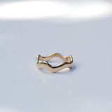 A gold wavy band ring with a polished finish, resting on a light gray surface with soft, artistic shadows.