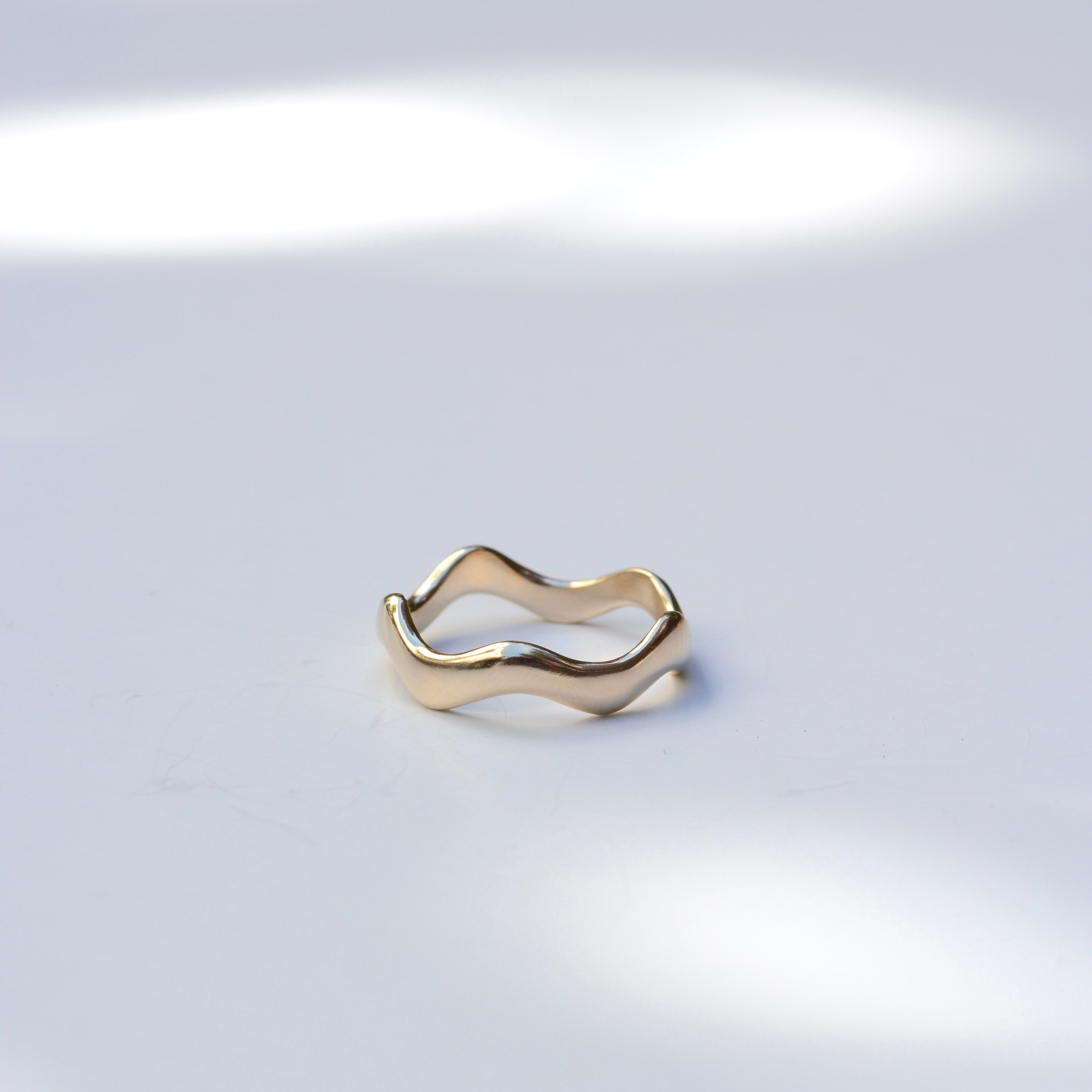 A slim, wavy open-band ring in polished yellow gold, photographed on a light gray surface with soft, dappled light shadows.