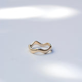 A slim, wavy open-band ring in polished yellow gold, photographed on a light gray surface with soft, dappled light shadows.