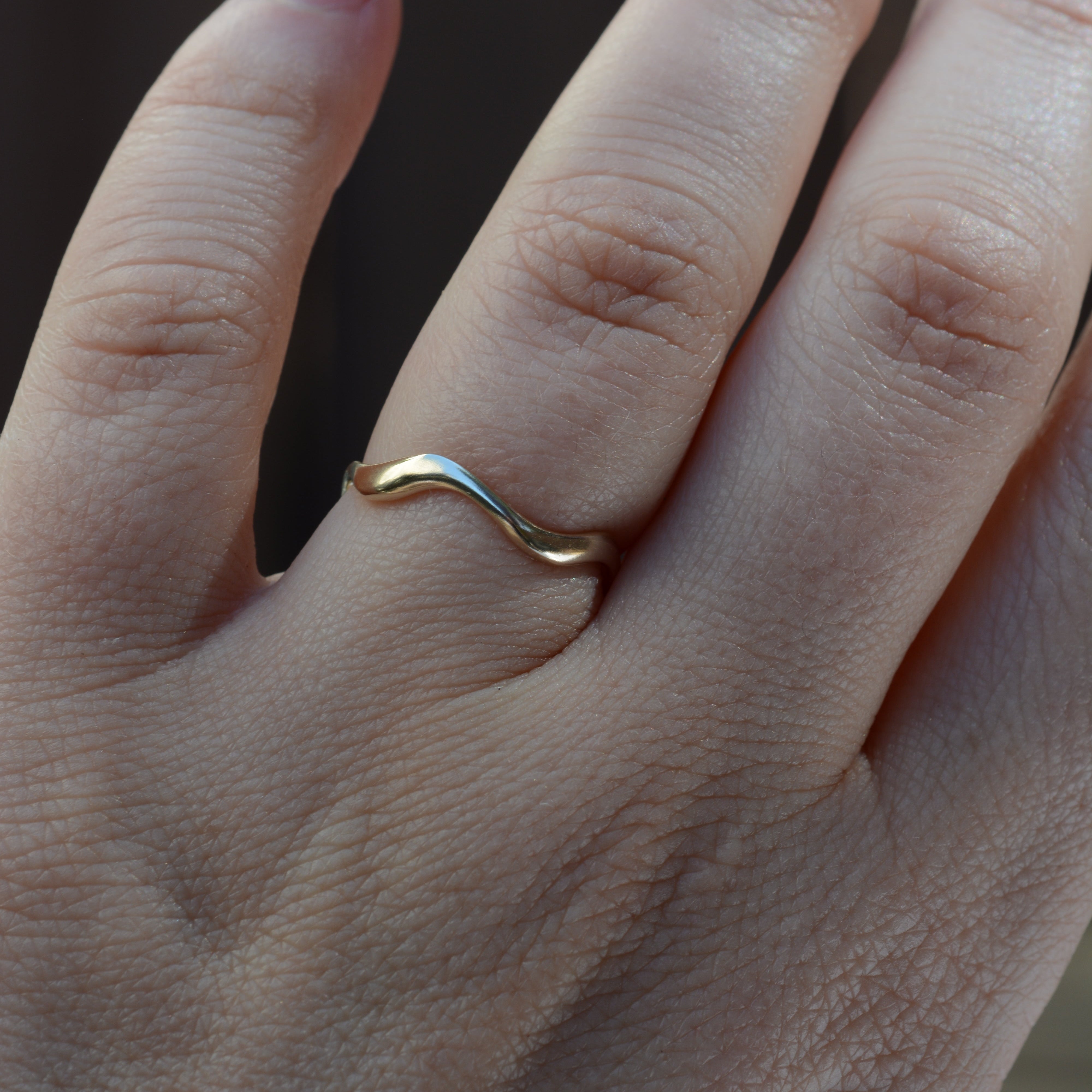 A close-up of a hand wearing a thin, wavy two-tone ring with gold and silver metals, set against a dark, blurred background.