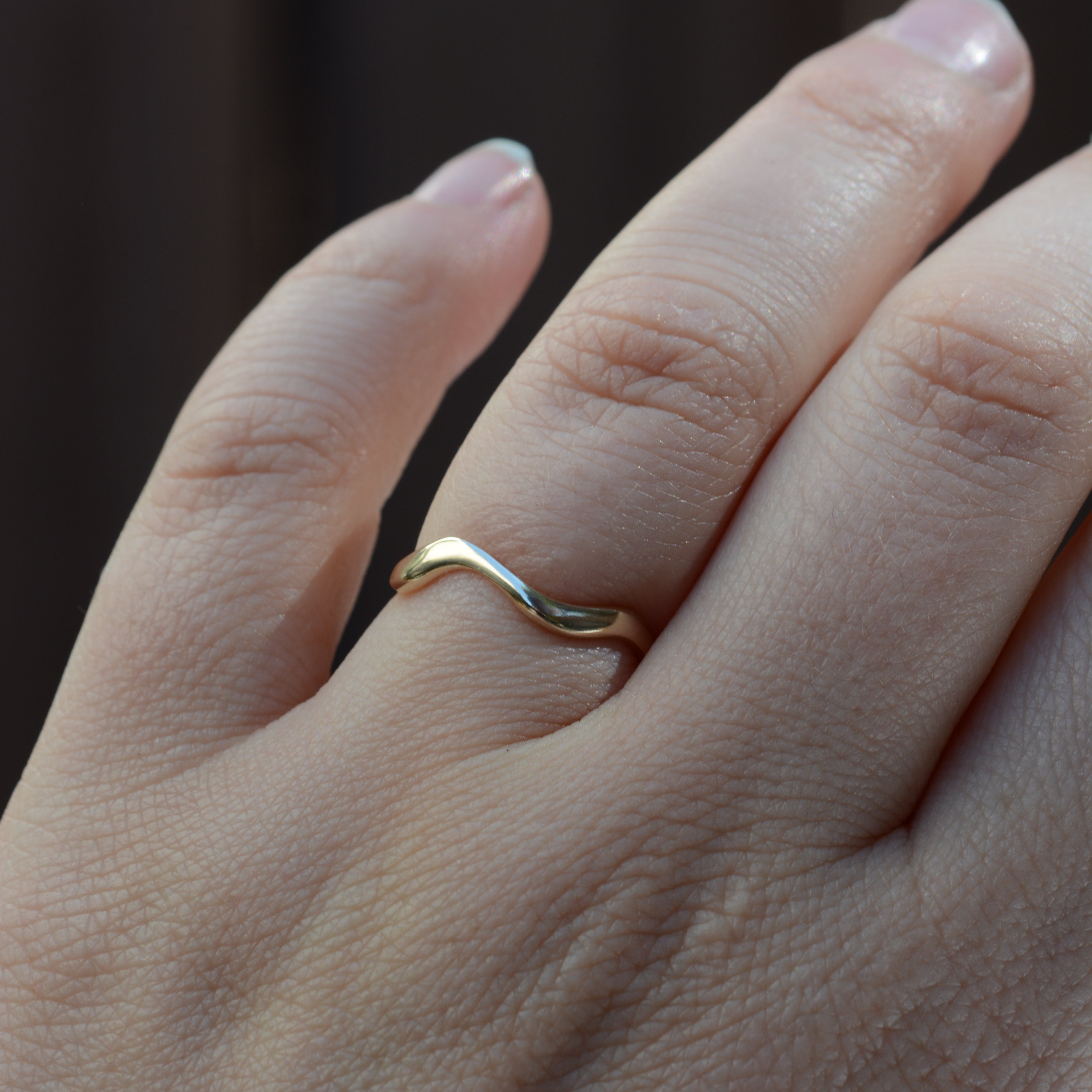 A close-up of a hand wearing a simple wavy band ring in yellow gold, set against a dark, blurred background.