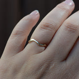 A close-up of a hand wearing a simple wavy band ring in yellow gold, set against a dark, blurred background.