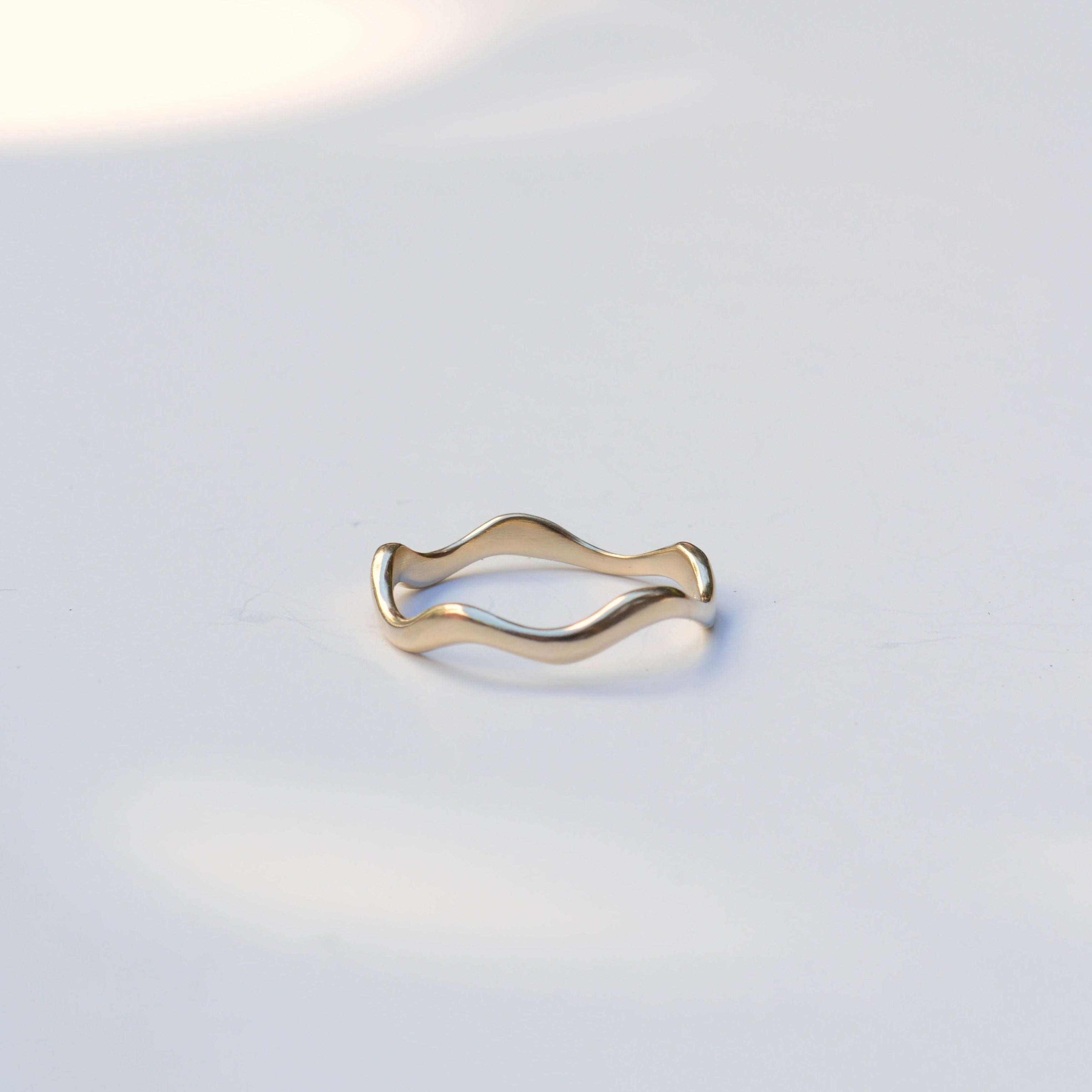 A thin wavy gold band ring resting on a light gray surface with soft shadows.