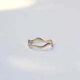 A thin wavy gold band ring resting on a light gray surface with soft shadows.
