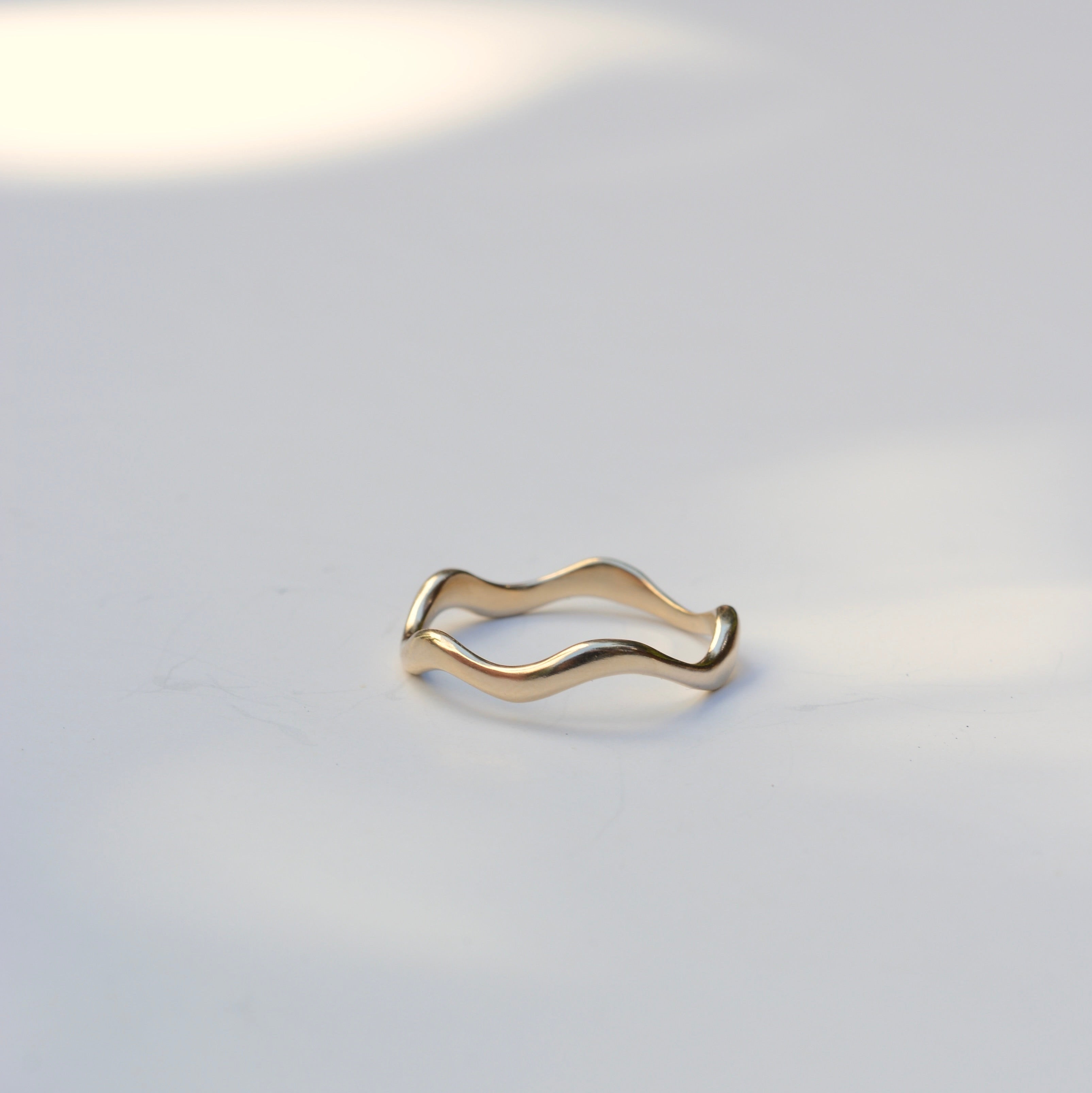 A thin, wavy gold ring with a polished finish, resting on a light gray surface with soft, natural shadows.
