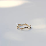 A thin, wavy gold ring with a polished finish, resting on a light gray surface with soft, natural shadows.