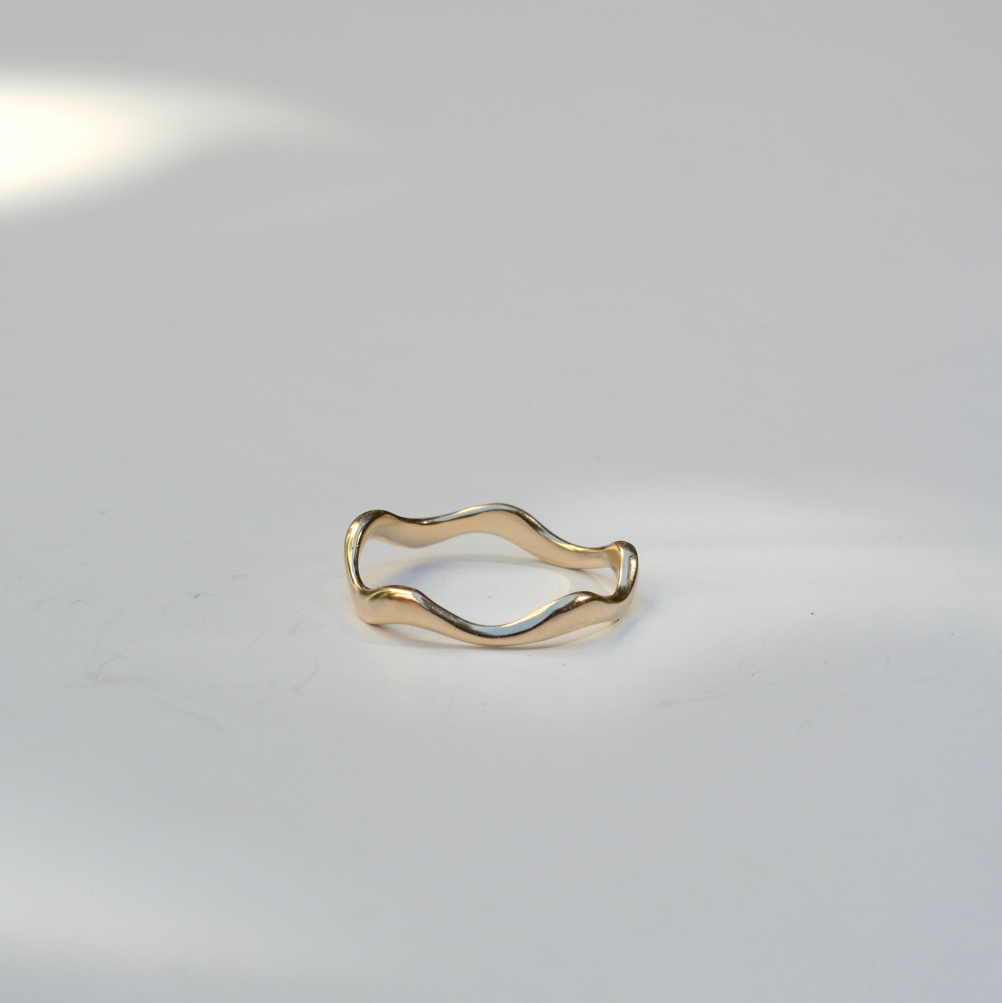 A thin, wavy gold ring with an organic, undulating shape, photographed on a clean white surface with soft shadows.