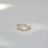 A thin, wavy gold ring with an organic, undulating shape, photographed on a clean white surface with soft shadows.