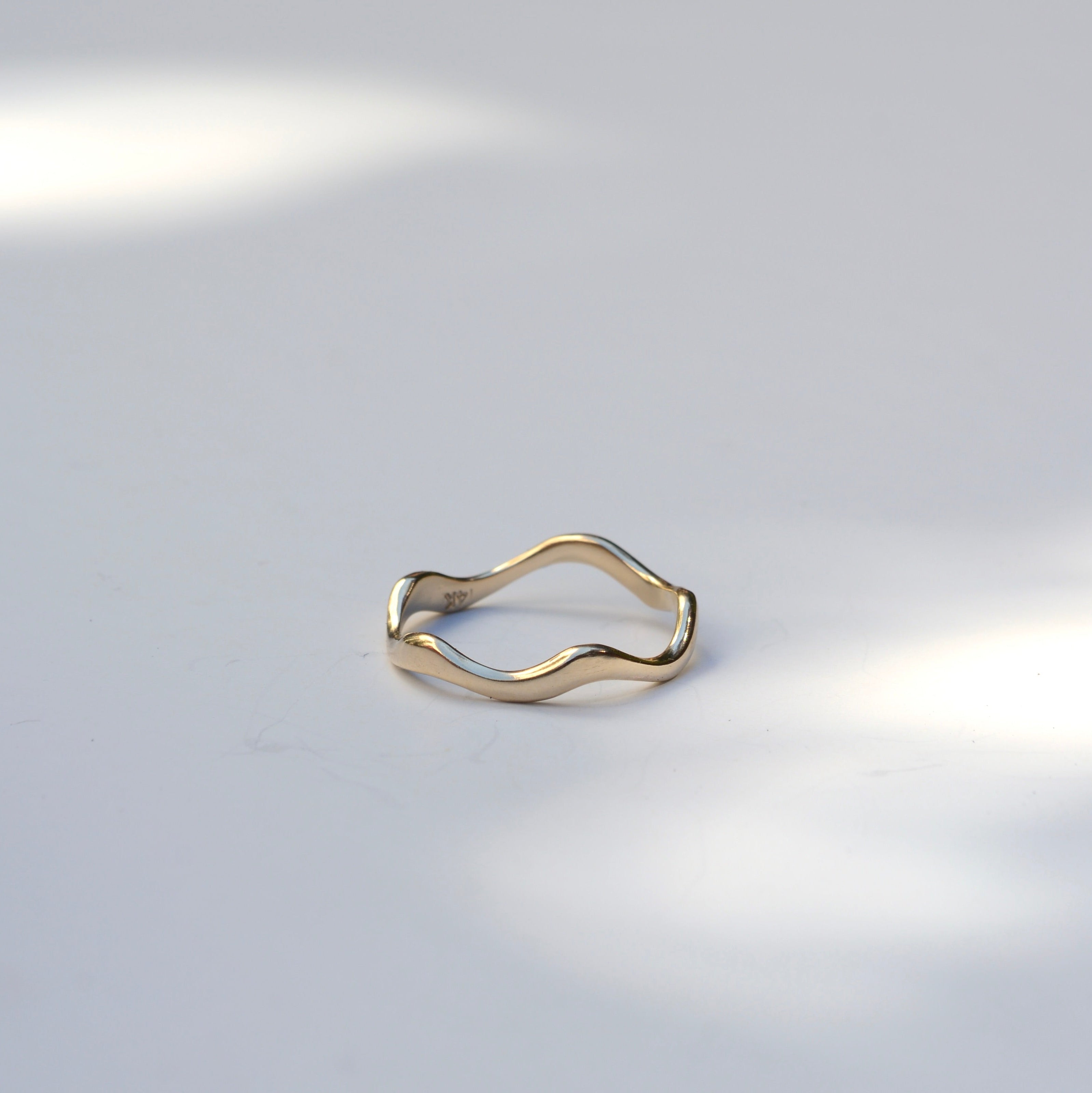 A thin, wavy open-band ring in polished yellow gold, photographed on a light gray surface with soft natural shadows.