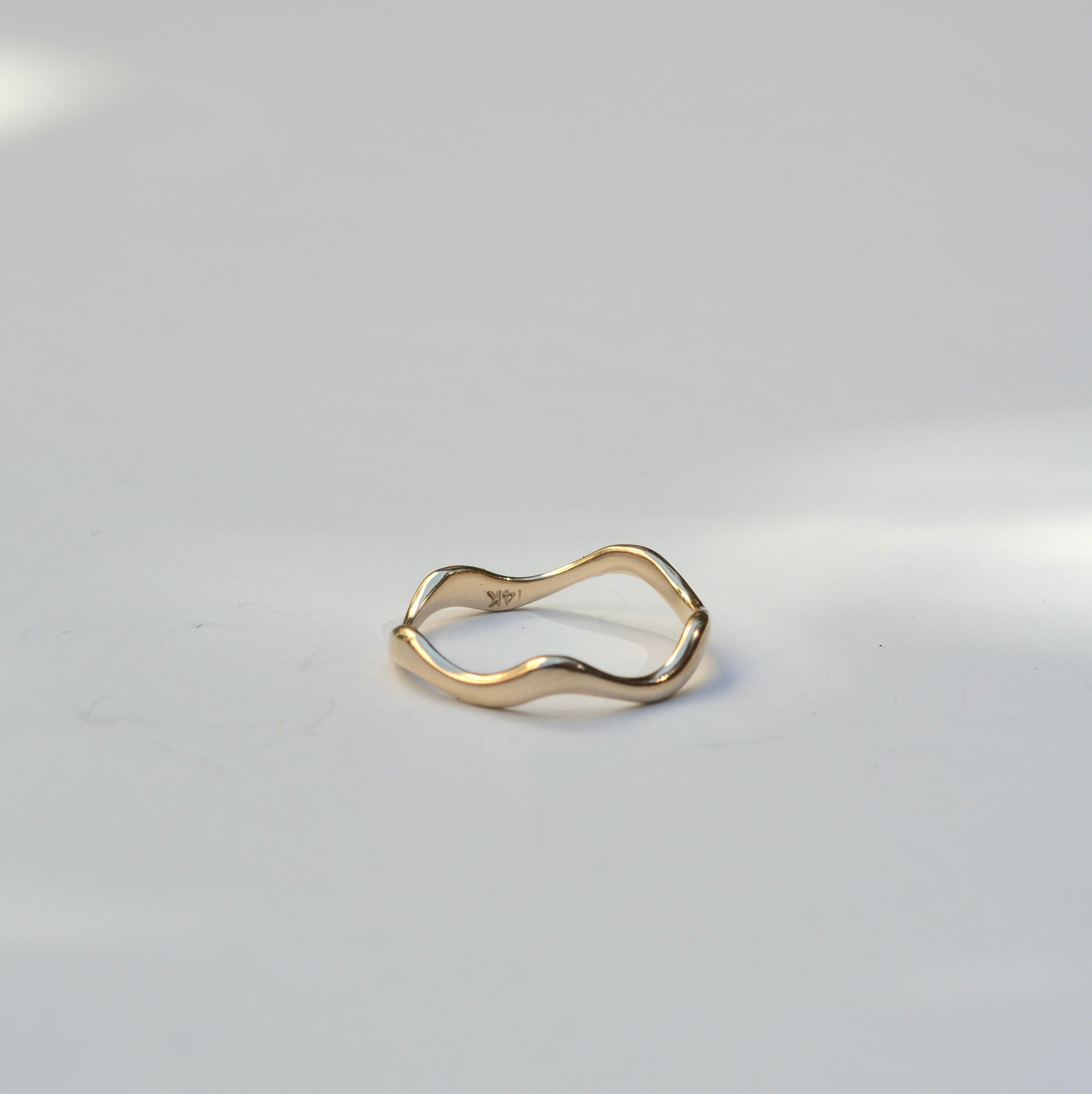 A single gold wavy band ring resting on a plain light gray surface.