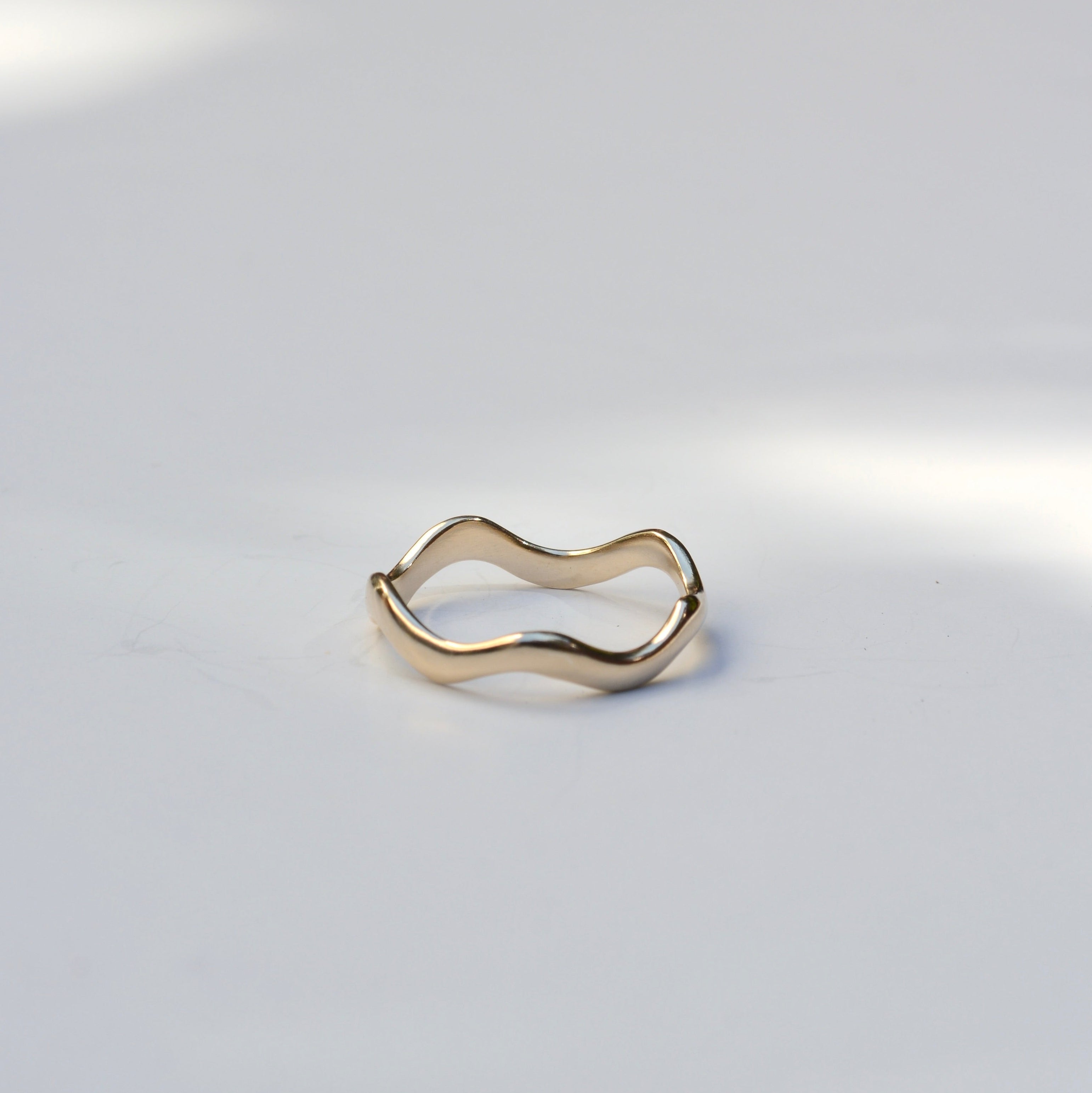 A slim, wavy open ring in polished yellow gold, photographed on a light gray surface with soft natural shadows.