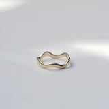 A slim, wavy open ring in polished yellow gold, photographed on a light gray surface with soft natural shadows.