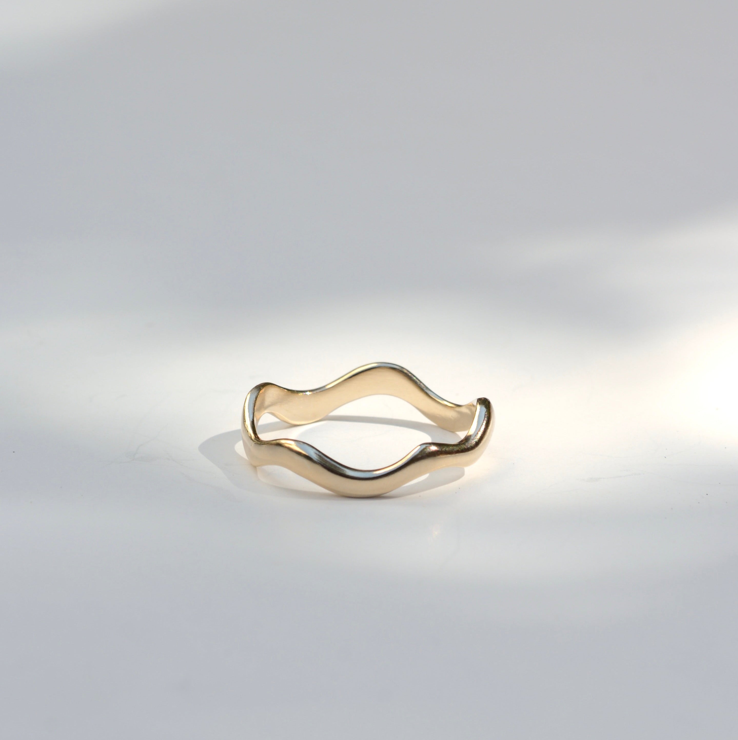 A thin wavy band ring in polished yellow gold, photographed on a light gray surface with soft shadows.