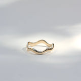 A thin wavy band ring in polished yellow gold, photographed on a light gray surface with soft shadows.
