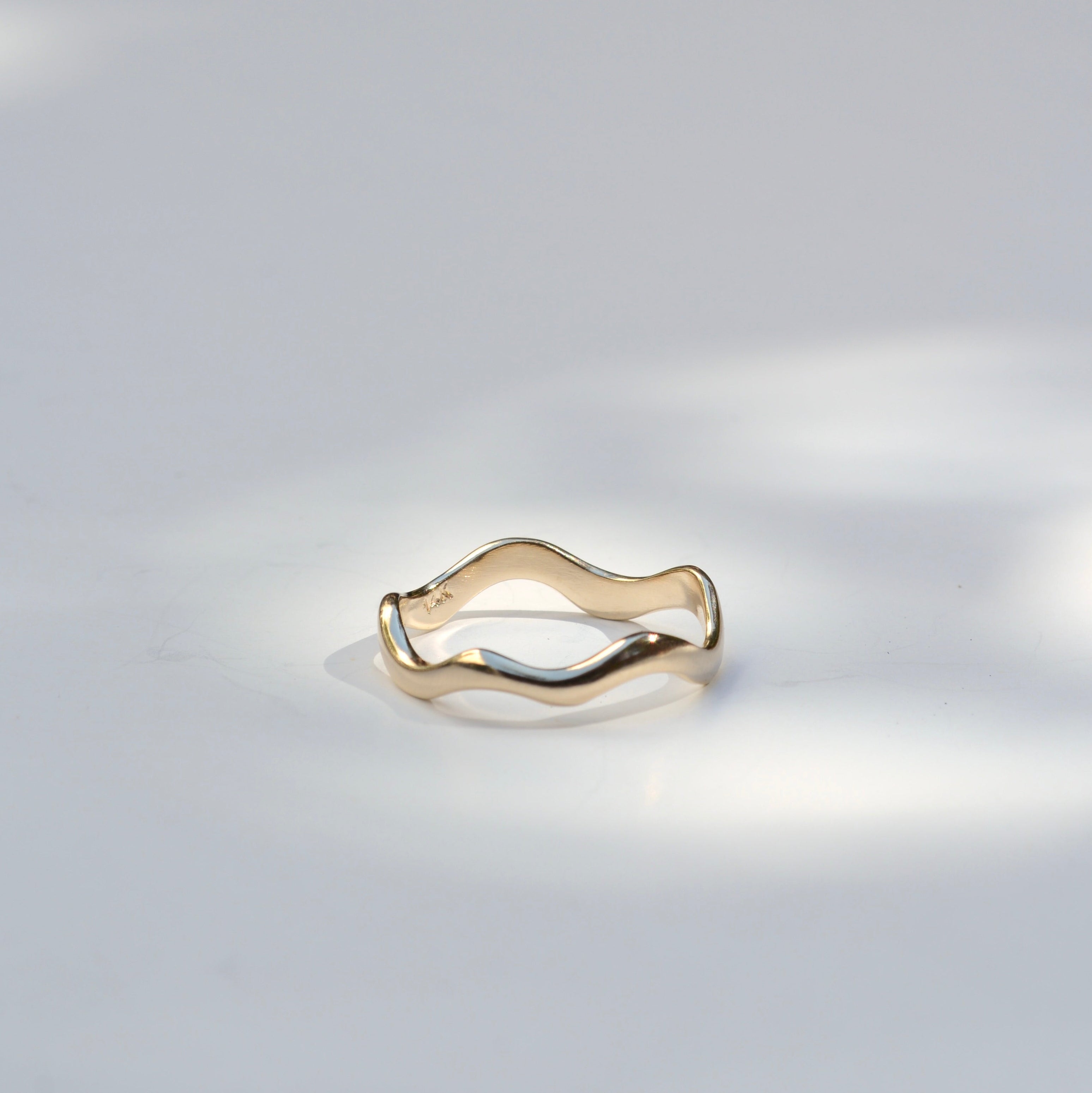 A thin wavy gold ring with a polished finish, resting on a light gray surface with soft, dappled shadows.