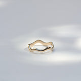 A thin wavy gold ring with a polished finish, resting on a light gray surface with soft, dappled shadows.