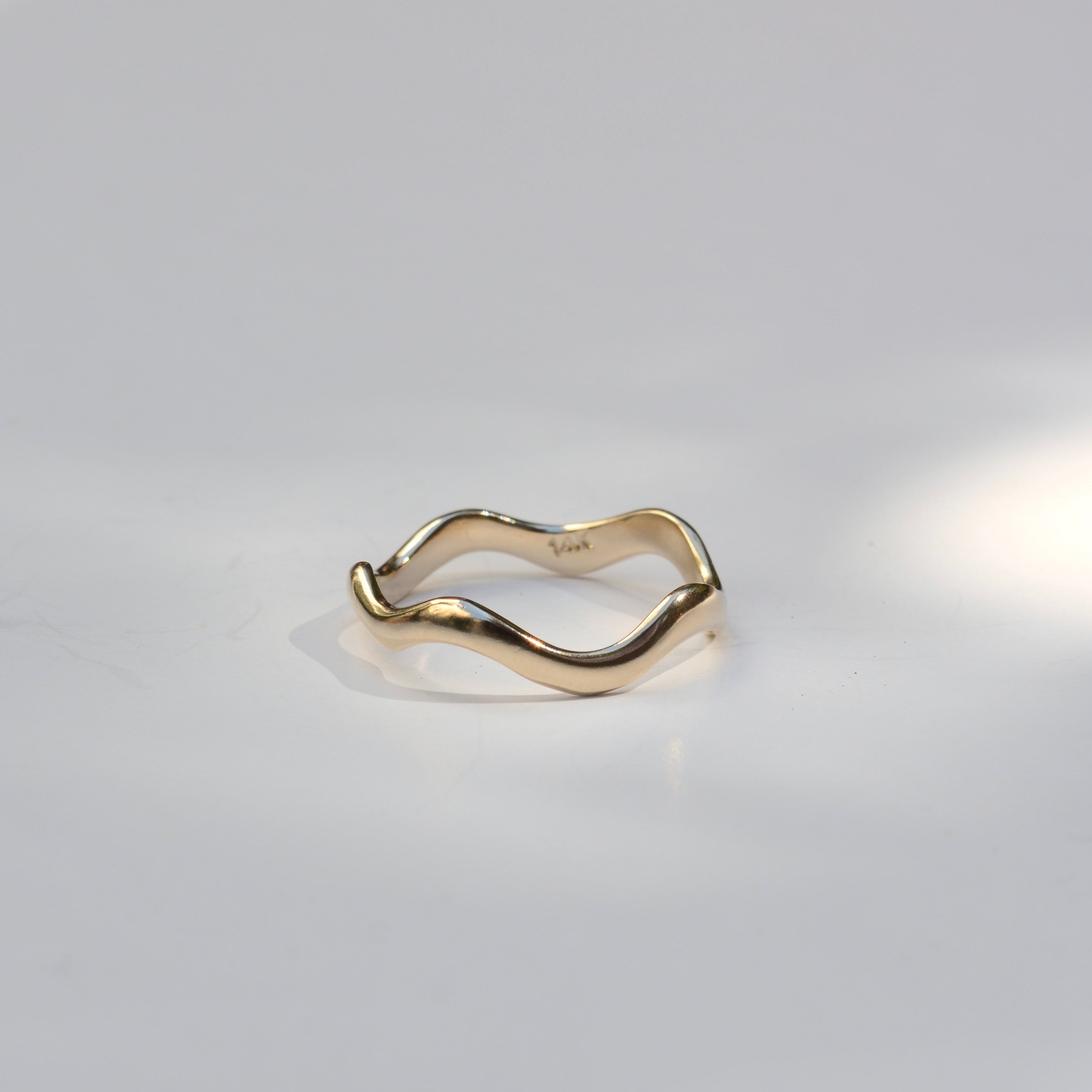 A single gold ring with a wavy, organic wavy band, photographed on a clean white surface with soft, artistic shadows.