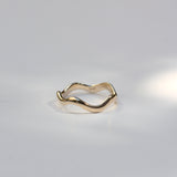 A single gold ring with a wavy, organic wavy band, photographed on a clean white surface with soft, artistic shadows.