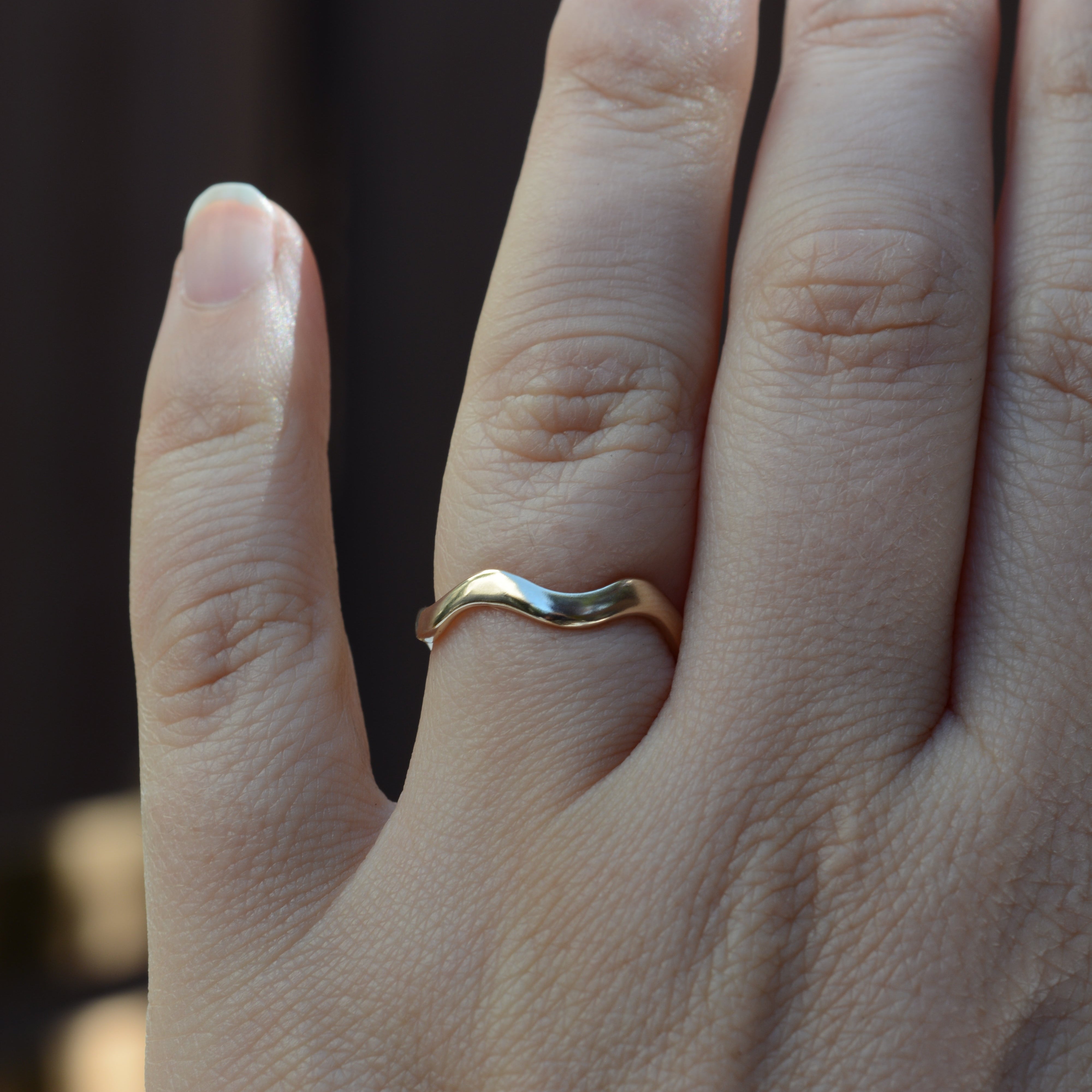 A close-up of a hand wearing a thin, wavy band ring in polished yellow gold, set against a dark, blurred background.