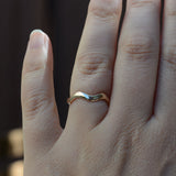 A close-up of a hand wearing a thin, wavy band ring in polished yellow gold, set against a dark, blurred background.