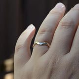 A close-up of a hand wearing a thin, wavy gold band ring against a dark, blurred background.
