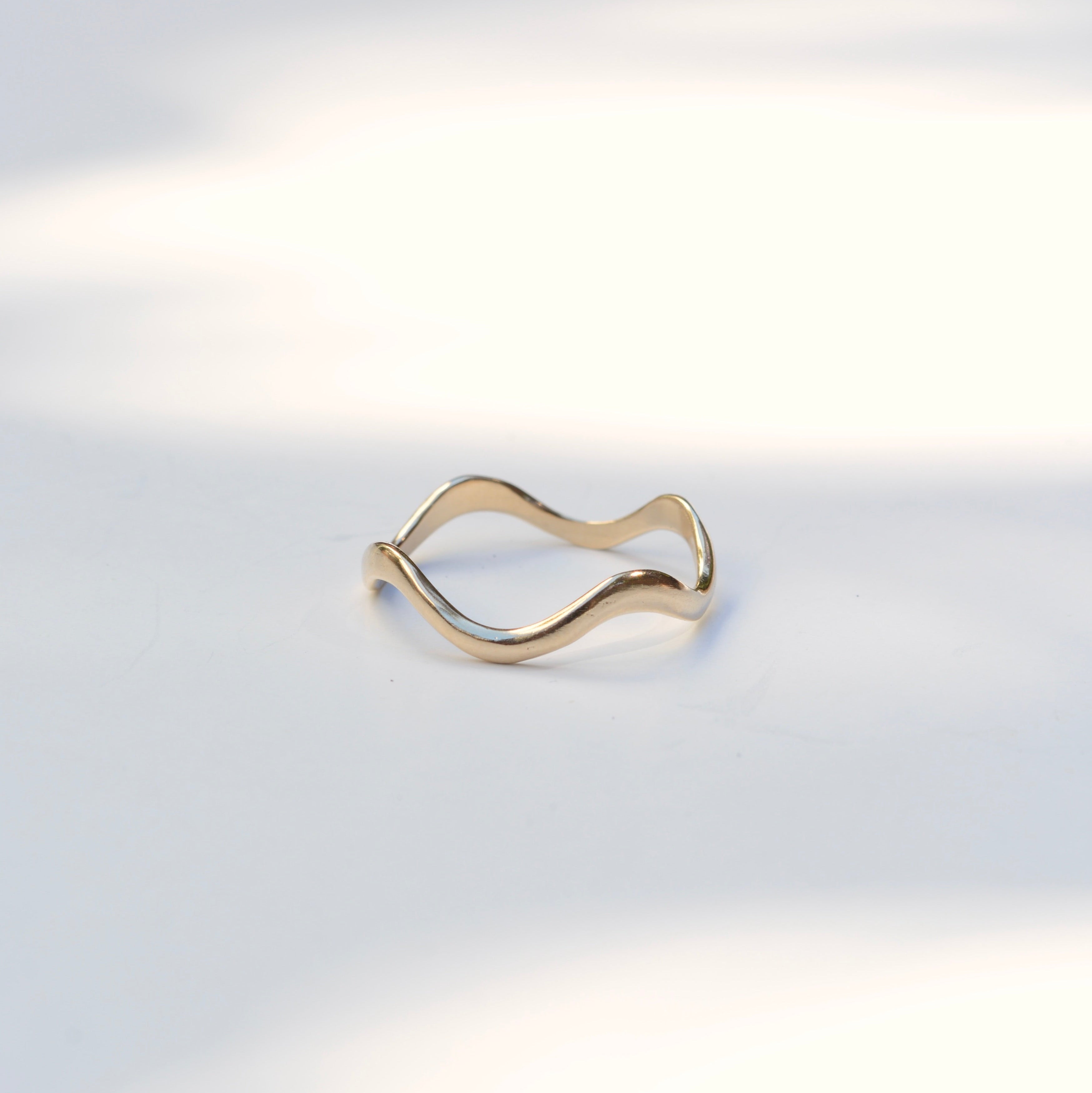A thin, wavy gold ring with a polished finish, displayed on a clean white surface with soft, natural shadows.